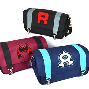 以下が含まれることがあります： 黒、バーガンディ、ネイビーブルーのメッセンジャーバッグ3つ。各バッグは、赤い「R」、赤いエンブレム、白と青のグラフィックというユニークなデザインが特徴です。バッグには調節可能なストラップとシルバーの金具が付いています。