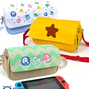 Könnte beinhalten: Drei farbenfrohe Umhängetaschen mit gestickten Designs, darunter eine blaue Tasche mit verschiedenen Symbolen, eine gelbe Tasche mit einem braunen Stern und eine grün-beige Tasche mit Blatt- und Fruchtmotiven. Unten befindet sich eine Nintendo Switch-Konsole.