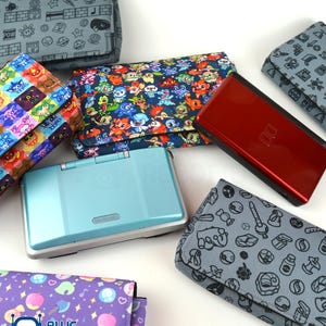 May include: A collection of Nintendo DS consoles and patterned cases. The cases feature various designs, including cartoon characters and video game-themed graphics. A light blue Nintendo DS is visible, along with a red one.
