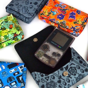 Puede incluir: Una colección de carteras coloridas y estampadas con una consola Game Boy Color. Las carteras presentan varios diseños, incluyendo personajes de videojuegos y patrones de controladores. Las carteras son en tonos de azul, naranja, verde y gris, con una cartera negra abierta para revelar la consola.