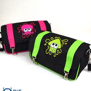 May include: Two black messenger bags with cartoon squid designs. One bag has a pink squid and pink straps, the other has a green squid and green straps. Both bags have silver hardware and adjustable straps.
