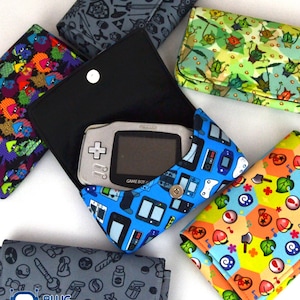 May include: A collection of colorful fabric wallets, each featuring unique patterns and designs. One wallet is open, revealing a Game Boy Advance. The wallets showcase various themes, including video game consoles, and other playful motifs. The wallets are closed with a snap.
