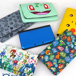May include: A collection of Nintendo 3DS cases and a blue 3DS console. The cases feature various designs, including a green Bulbasaur face, a yellow Pikachu face, and other colorful character patterns. The cases are designed to protect the console.