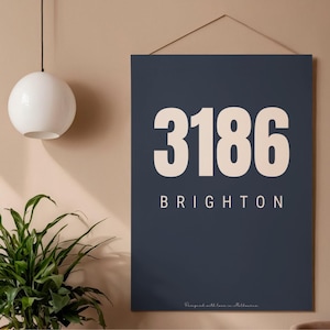 May include: A navy blue wall art print featuring the number "3186" in large white font above the word "BRIGHTON". The print is hanging on a wall with a neutral color scheme. A white spherical light fixture and a green plant are in the background.