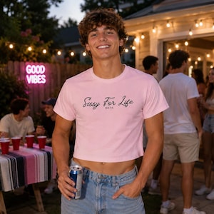May include: A light pink crop top with the text "Sissy For Life BETA" in black script. The shirt is worn by a person in blue jeans, holding a can of soda. The background includes a "Good Vibes" neon sign and a party setting.