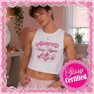 Puede incluir: Una camiseta corta sin mangas blanca con el texto rosa Addicted To Sissy Hypno e ilustraciones gráficas de un plug, un corazón, ropa interior y un espejo compacto. La prenda se combina con pantalones cortos rosa claro y un collar con corazón.