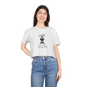 Puede incluir: Camiseta blanca corta con la palabra "SISSY" encima de un gráfico de una persona arrodillada y la palabra "SLAVE" debajo. El gráfico es en negro y rojo. La modelo lleva vaqueros azules.