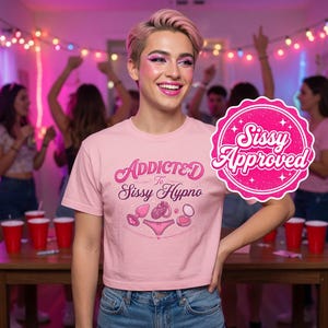 May include: A light pink short-sleeved t-shirt featuring the text Addicted To Sissy Hypno and graphic illustrations of a plug, a ring, a pair of panties, and a compact. A pink circular badge with the text Sissy Approved is on the right side.
