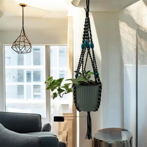 May include: A hanging plant holder made of braided black cord with teal accent beads, suspending a sage green ribbed ceramic pot containing a green ivy plant. The macrame structure features a knotted design with a tassel at the bottom.