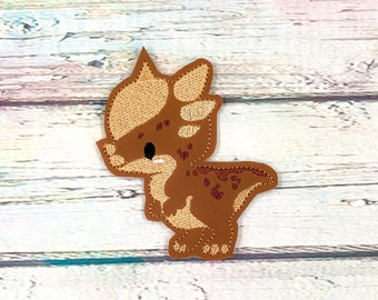 May include: A brown and tan dinosaur patch with embroidered details. The patch features a cartoon-style dinosaur with a spiky head and a spotted pattern on its body. The patch is likely for clothing or accessories.