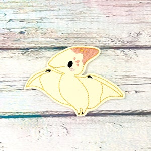 May include: An embroidered patch featuring a cartoon pterodactyl with a cream-colored body, pink accents, and black eyes. The patch has gold stitching around the edges, giving it a polished look. The design is set against a light-colored wooden background.
