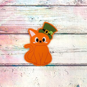May include: Orange cat patch wearing a green leprechaun hat with a black buckle and shamrock. The cat has white accents on its face and ears, and a stitched outline. The patch is on a light wood grain background.