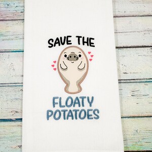 May include: White hand towel with embroidered text and a cartoon manatee design. The text reads "SAVE THE" above the manatee and "FLOATY POTATOES" below. The manatee is beige with black eyes and a smiling face, surrounded by small red hearts.