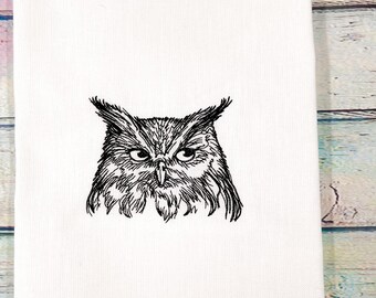 May include: White kitchen towel featuring a detailed black embroidered owl design. The owl's face is centered, showcasing intricate feather details and piercing eyes. The towel is made of a soft, absorbent material, perfect for everyday use.