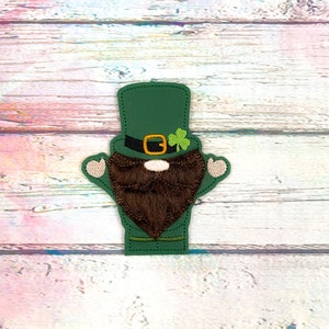 May include: A green leprechaun-themed applique with a top hat, beard, and shamrock. The hat has a black band with a gold buckle. The beard is brown, and the shamrock is green. The applique has small beige hands.