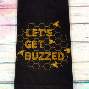 May include: Black kitchen towel with gold embroidered text and bee-themed design. The text reads "LET'S GET BUZZED" with bees and honeycomb patterns. A decorative item for the kitchen.
