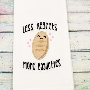May include: White kitchen towel with the embroidered text "Less Regrets More Baguettes". The towel features a smiling baguette with pink heart accents. The design is in black and tan.