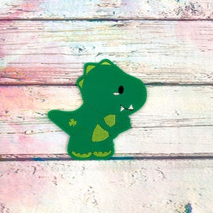 May include: A green dinosaur applique with a shamrock detail. The dinosaur has a friendly expression with a white tooth and a black eye. The applique is made of a smooth material and has a stitched outline.