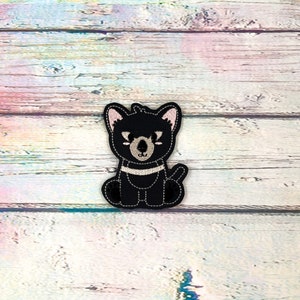 May include: A black embroidered patch featuring a cartoon Tasmanian devil. The patch has white stitching details, pink inner ears, and a white stripe across the chest. The background is a light-colored wooden surface.