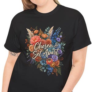 Peut inclure: Un t-shirt noir à manches courtes orné d'un motif floral coloré composé de fleurs orange, bleues et violettes. Le texte "Chosen & Set Apart" est imprimé en écriture blanche au centre du bouquet, avec "1 PETER 2:9" inscrit en dessous.
