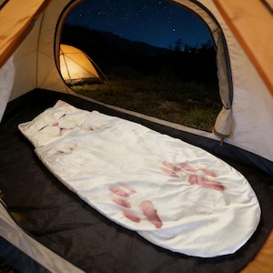 May include: A white cat-shaped sleeping bag with pink paw prints inside a tent. The sleeping bag has a cat face design on the pillow area. Another tent is visible in the background under a starry night sky.