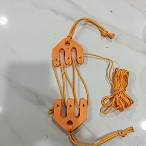 May include: An orange pulley system with yellow rope. The pulley has two orange, bracket-shaped pieces connected by the rope. The rope is knotted at the ends and is also coiled. The pulley system is designed for lifting or pulling objects.