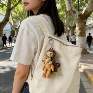 May include: A brown and tan monkey keychain with a silver keyring attached to a white canvas tote bag. The keychain features two monkey figurines, one hugging the other. The background shows a street with trees and people.