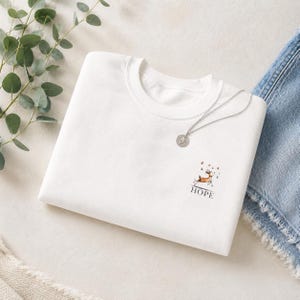 May include: A white crew neck sweatshirt with a small deer graphic and the word "HOPE" printed below it. A silver necklace with a round pendant rests on the shirt. Denim jeans are visible in the background.