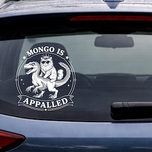 Può includere: Adesivo in vinile bianco raffigurante un gatto con occhiali da sole e una corona che cavalca un dinosauro. Il testo "MONGO IS APPALLED" è visualizzato in un design circolare con stelle e un banner.