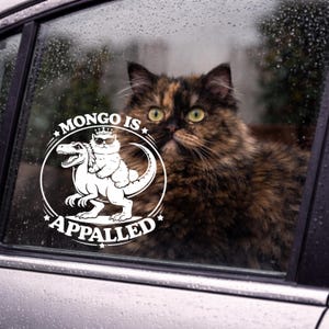 May include: A car window sticker with a white graphic of a cat wearing sunglasses and a crown, riding a dinosaur. The text "MONGO IS APPALLED" is in a circle around the image. A tortoiseshell cat is visible through the window.