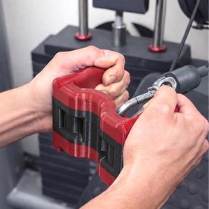 May include: Red and black weight machine handle. The handle is attached to a cable with a metal clip. The handle is being held by a person. The handle is red with black accents.