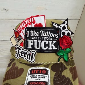 Könnte beinhalten: Eine Camouflage-Trucker-Mütze mit verschiedenen Aufnähern. Zu den Aufnähern gehören Spielkarten, eine rote Rose, ein schwarz-weißer Stern und ein Aufnäher mit der Aufschrift "I like Tattoos AND THE WORD FUCK". Auf der Mütze steht auch das Wort "Feral".