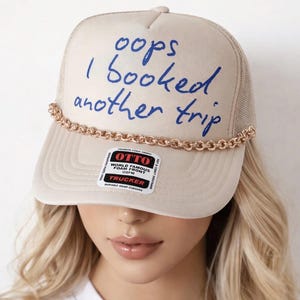 May include: A beige trucker hat featuring a mesh back and a foam front with the blue text "oops I booked another trip" printed on it. A gold-toned chain trim runs across the base of the crown, and a rectangular sticker is on the brim.