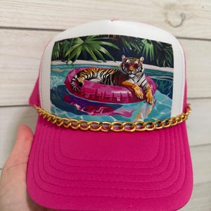 May include: A bright pink trucker hat featuring a white front panel with a graphic print of a tiger wearing sunglasses while floating on a pink inner tube in a swimming pool. A gold-colored metal chain is attached across the base of the front panel.