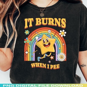 May include: Black t-shirt with a retro design featuring a yellow star character, a rainbow, and the text "IT BURNS WHEN I PEE". The design includes flowers and stars.