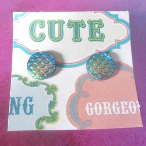 May include: A pair of blue and green iridescent mermaid scale stud earrings on a white card with the word "CUTE" in green and blue letters and the words "NG" and "GORGEO" in black letters.