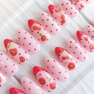 Può includere: Un set di unghie finte a forma di mandorla. Ogni unghia è dipinta di rosa chiaro con piccoli cuori rossi e un disegno di ciliegia. Le punte sono dipinte di rosso e i disegni delle ciliegie variano.