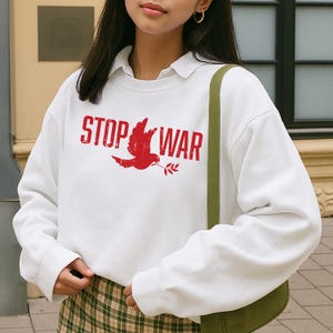 May include: White sweatshirt with the red text "STOP WAR" and a red dove carrying an olive branch. The sweatshirt has a collared neckline and long sleeves. The person is wearing a green bag.