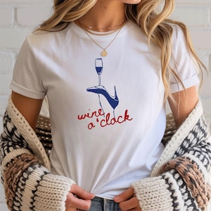 May include: White t-shirt with a graphic of a champagne glass, a high-heeled shoe, and the words "wine o'clock" in red and blue. The shirt is made of a soft material.