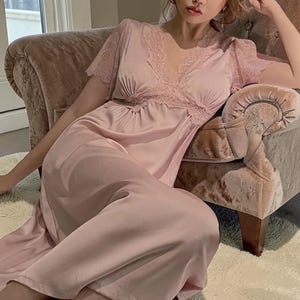May include: A light pink satin nightgown with lace trim around the neckline and sleeves. The V-neck nightgown is a soft, flowing design. The model is sitting in a pink velvet chair.