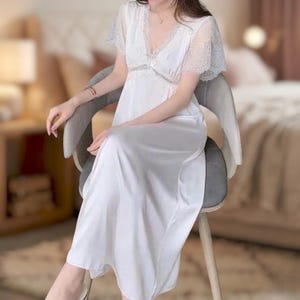 May include: A white satin nightgown with lace detailing on the sleeves and neckline. The dress has a V-neck and a flowing, floor-length skirt. The model is seated on a gray chair. The background is blurred, suggesting a bedroom setting.