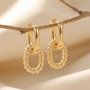 May include: A pair of gold-colored hoop earrings. Each earring features a twisted oval pendant suspended from a simple hoop. The earrings are displayed on a thin, brown branch against a soft, neutral background.