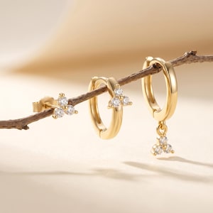 May include: A set of gold earrings with a floral design. The earrings are displayed on a small branch. One earring is a stud, and the other two are hoop earrings with a dangling floral charm. Each floral design has three clear, round gemstones.