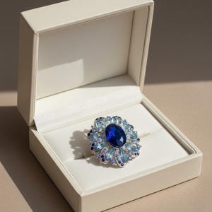 May include: A floral ring featuring a large, oval, deep blue gemstone at its centre, encircled by smaller light blue and dark blue stones. The ring is set in silver and displayed in an open, off-white jewellery box.
