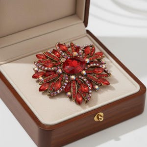 Peut inclure: Une broche rouge ornée, de forme florale, présentée dans un écrin en bois. La broche est ornée d'une grosse pierre rouge centrale, entourée de petits cristaux rouges et irisés. L'intérieur de la boîte est de couleur claire.