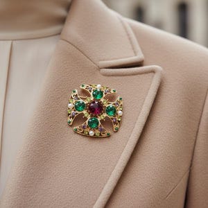 Peut inclure: Broche dorée en forme de croix, ornée d'une pierre rouge au centre, entourée de pierres vertes et de perles. La broche est fixée sur un blazer beige, un accessoire de mode classique.