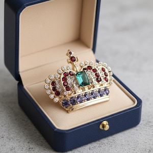 Peut inclure: Broche couronne dorée ornée de pierres précieuses rouges, transparentes, vertes et violettes, présentée dans un écrin bleu à intérieur beige. La broche est très détaillée et ornée.