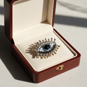Peut inclure: Broche dorée en forme d'œil avec une pierre centrale bleu foncé, entourée de cristaux blancs et transparents. La broche est présentée dans un écrin à bijoux en bois foncé ouvert, à l'intérieur crème.