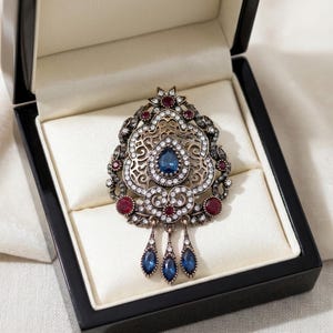 Peut inclure: Broche ornée avec un motif en filigrane, sertie de pierres précieuses bleues et rouges et de cristaux transparents. La broche est présentée dans un écrin noir avec un intérieur crème. La broche a trois pendentifs bleus en forme de goutte.