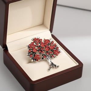 Peut inclure: Broche argentée en forme d'arbre ornée de cristaux rouges, présentée dans un écrin marron à intérieur crème. La broche présente un motif d'arbre détaillé avec des 'feuilles' en cristal rouge et un tronc texturé.